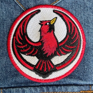 May include: Embroidered patch featuring a red cardinal bird with black wings, set against a white background within a red-bordered circle. The patch is sewn onto a denim jacket, showcasing the bird's detailed design.