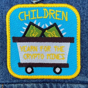 May include: A rectangular embroidered patch with a yellow border on a blue denim fabric. The patch features a light blue background with the text "CHILDREN" in yellow, and a mine cart graphic containing green shapes with yellow lines.