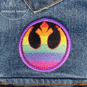 May include: Embroidered patch featuring a rainbow-colored design within a purple circular border. The patch is sewn onto a denim jacket. The design includes a black symbol with orange, yellow, green, blue, and pink hues.