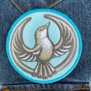 May include: A circular embroidered patch featuring a grey bird with outstretched wings centered on a light blue background. The bird has a white chest, a small yellow eye, and is framed by a bright cyan border, all stitched onto blue denim fabric.