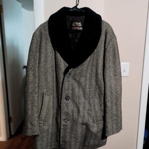 May include: A long coat featuring a grey and black herringbone pattern with a large black faux fur shawl collar. The coat has a three-button front closure, two side pockets, and a black interior lining with an Oakbrook Sportswear label.
