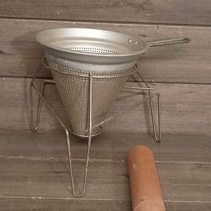 Pode incluir: Um passador cónico ou chinês de metal prateado com corpo perfurado, cabo comprido e base de suporte em arame, acompanhado por um pilão de madeira cilíndrico separado com uma extremidade arredondada para pressionar os alimentos.
