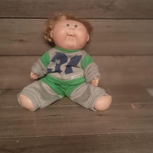 May include: A vintage Cabbage Patch Kids doll, featuring a round face, blonde hair, and blue eyes. The doll is wearing a green and gray outfit with the number 24 on the front. The doll is sitting on a wooden surface.