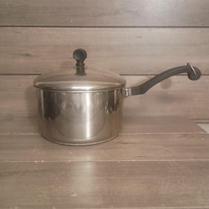 Può includere: Un pentolino in acciaio inossidabile con finitura lucida, dotato di coperchio coordinato con pomello nero e un lungo manico nero ricurvo. La pentola ha un corpo cilindrico e una base piatta, progettata per l'utilizzo sui fornelli.