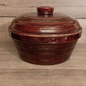 Puede incluir: Una fuente de cerámica de color rojo oscuro con tapa. El plato tiene forma redonda con un borde ligeramente acampanado y un patrón decorativo alrededor de la base. La tapa tiene un pequeño tirador de pomo.