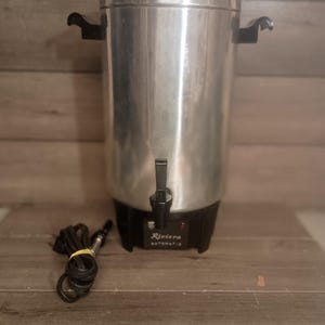 May include: A vintage Riviera automatic coffee urn with a stainless steel body and black base. It has a spigot, two black handles, and a black lid. The power cord is coiled and secured with a yellow band.