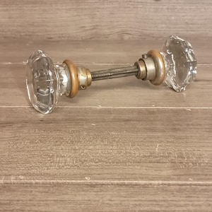 May include: Clear glass door knob with a faceted design. The knob has a brass-colored metal base and a threaded rod connecting the two knobs. The background is a light brown wooden surface.