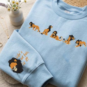 Puede incluir: Una sudadera azul claro de cuello redondo con una serie de cinco figuras de león bordadas en el pecho. La manga izquierda muestra una cabeza de león bordada con el texto "Life's not fair, is it?" cosido en hilo naranja sobre la cara.