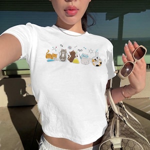 Peut inclure: Un t-shirt blanc à manches courtes orné d'un imprimé graphique représentant cinq chats de dessin animé interagissant avec de la verrerie de laboratoire et des symboles scientifiques, incluant le texte O2 et H2O, un atome et des béchers.