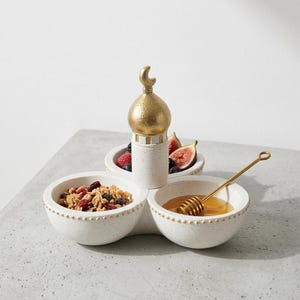 Könnte beinhalten: Ein weißes Keramik-Serviertablett mit drei Abschnitten, die jeweils Lebensmittel enthalten. Ein goldfarbenes, moscheeförmiges Mittelstück sitzt oben auf dem Tablett. Das Tablett enthält Müsli, Honig und Obst. Ein goldener Honiglöffel liegt im Honig.