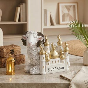 Peut inclure: Une décoration blanche et dorée pour l'Aïd al-Adha représentant un mouton et une mosquée. Le mouton a une laine blanche bouclée et des cornes brun foncé. La mosquée a des dômes dorés et des croissants de lune. Les mots "Eid al Adha" sont écrits sur la base.