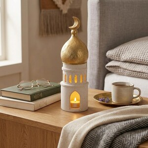Peut inclure: Un bougeoir décoratif en forme de mosquée avec un dôme doré et un croissant de lune. La base blanche présente des fenêtres en arc et une bougie allumée à l'intérieur. Une tasse de thé, des livres et des lunettes sont à proximité.