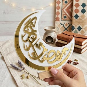 Peut inclure: Décoration en forme de croissant de lune blanc avec une bordure dorée et des calligraphies arabes. La lune est maintenue par une petite base en bois. L'arrière-plan comprend un petit diffuseur d'huile, une pile de journaux en cuir et du tissu décoratif.