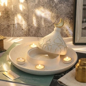 May include: A speckled white ceramic candle holder with a mosque-shaped lid and a gold crescent moon finial. The holder has a circular base with spaces for tea light candles. The lid is decorated with mosque details.