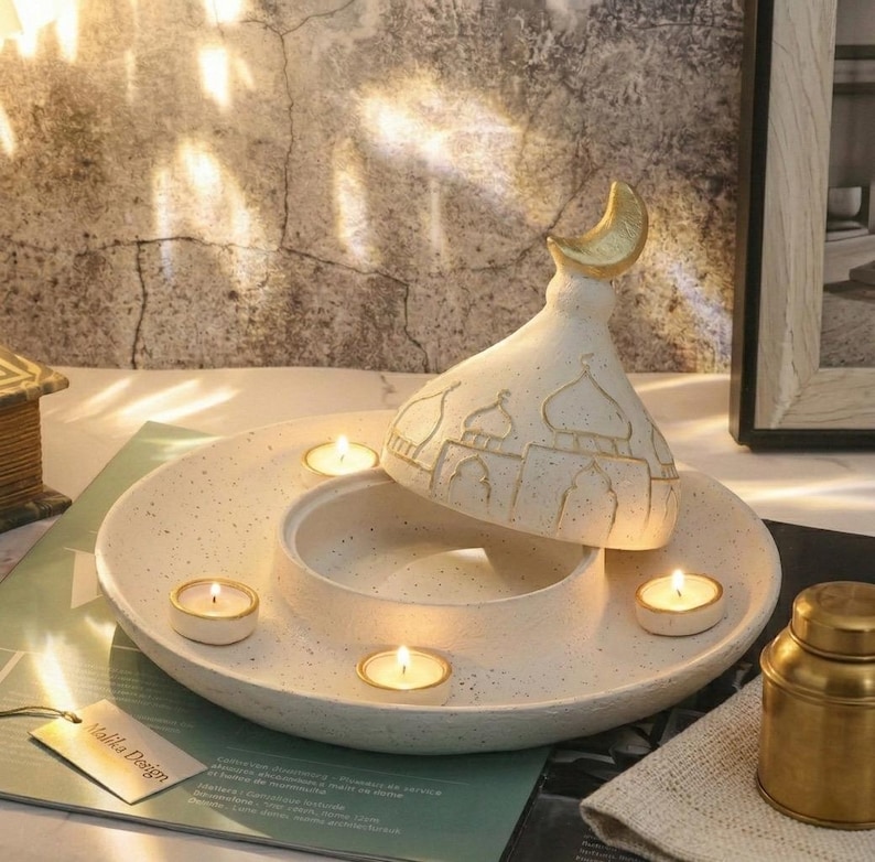May include: A speckled white ceramic candle holder with a mosque-shaped lid and a gold crescent moon finial. The holder has a circular base with spaces for tea light candles. The lid is decorated with mosque details.