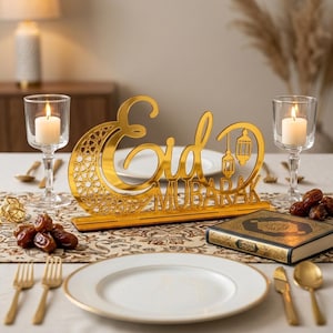 Może przedstawiać: Złota dekoracja stołu "Eid Mubarak" z motywem półksiężyca i lampionów. Na stole znajdują się zapalone świece, talerze, sztućce, daktyle i książka, tworząc świąteczną atmosferę.