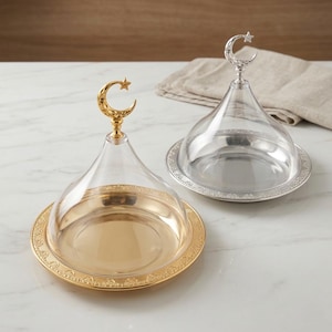 May include: Two decorative serving dishes with clear lids. One dish is gold-toned with a crescent moon and star handle, the other is silver-toned. Both have ornate rims and are on a white marble surface.