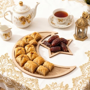 Peut inclure: Une table joliment dressée avec une théière blanche, une tasse et un pot à crème, ornés de motifs floraux dorés. Un plateau en bois en forme de croissant de lune contient des baklavas et des dattes. Une lanterne décorative et une nappe blanche complètent la scène.