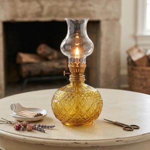 Può includere: Una lampada a olio in stile vintage accesa, con un camino in vetro trasparente e una base in vetro giallo a forma di diamante. La lampada è su un tavolo bianco con un piattino di fiammiferi, fiori secchi e piccole forbici.