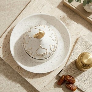 May include: A white ceramic serving dish with a lid featuring a gold mosque design and a gold handle. The dish sits on a speckled white plate. A small gold container and dates are also present.