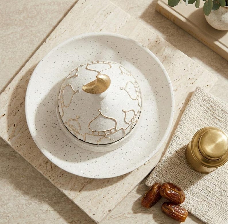 May include: A white ceramic serving dish with a lid featuring a gold mosque design and a gold handle. The dish sits on a speckled white plate. A small gold container and dates are also present.