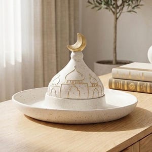 Può includere: Un elemento decorativo in ceramica bianca maculata con un coperchio a forma di moschea e un terminale a mezzaluna dorata. Il pezzo poggia su un vassoio maculato abbinato, adatto all'arredamento della casa.