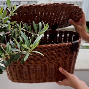 Può includere: Un cesto intrecciato marrone con coperchio incernierato e chiusura con cinturino in pelle marrone scuro. Il cesto presenta una forma rettangolare con angoli arrotondati ed è posizionato accanto a un ramo d'ulivo con foglie strette.