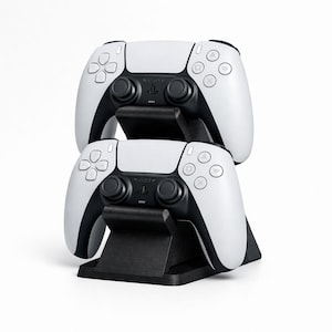 Puede incluir: Dos mandos de juego blancos y negros apilados sobre un soporte negro. Los mandos tienen un diseño simétrico con botones y pads direccionales. El soporte es rectangular con una ranura para sujetar cada mando.