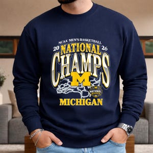 May include: Navy blue sweatshirt with the text "NCAA Men's Basketball 20 National Champs 26 Michigan". The design includes a yellow "M" and a basketball net graphic. The sweatshirt is a crew neck style.