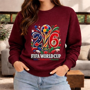 May include: Burgundy sweatshirt featuring a colorful graphic for the 2026 FIFA World Cup. The design includes a soccer ball, the numbers 2 and 6, and the flags of USA, Canada, and Mexico. The text "FIFA WORLD CUP" is displayed below.