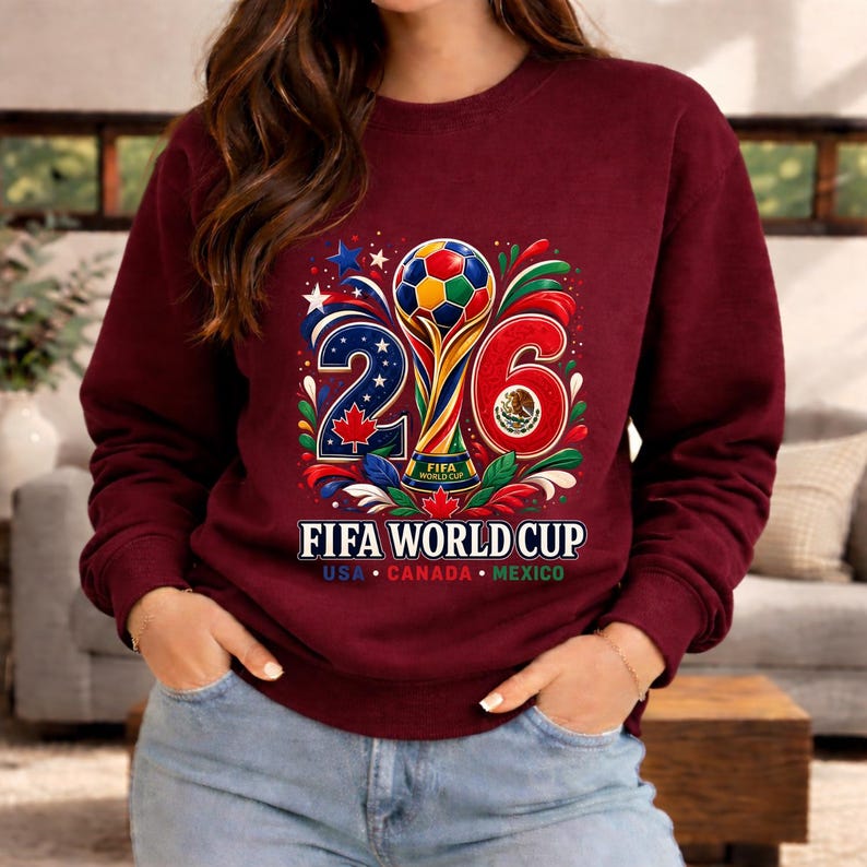 May include: Burgundy sweatshirt featuring a colorful graphic for the 2026 FIFA World Cup. The design includes a soccer ball, the numbers 2 and 6, and the flags of USA, Canada, and Mexico. The text "FIFA WORLD CUP" is displayed below.