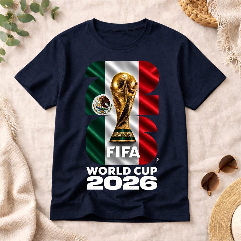 Pu&ograve; includere: Una t-shirt blu navy con una grande grafica del trofeo della Coppa del Mondo FIFA 2026 centrata sulla bandiera messicana. Il design include il testo FIFA WORLD CUP 2026 in lettere bianche, con il numero 2026 integrato nello sfondo della bandiera.