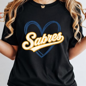 Peut inclure: T-shirt noir avec un cœur bleu et le mot "Sabres" en police rétro. Le texte est blanc avec un contour jaune. Un t-shirt décontracté, idéal pour les fans.