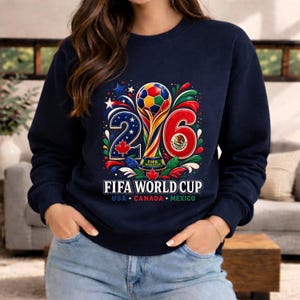 May include: Navy blue sweatshirt featuring a colorful graphic for the FIFA World Cup 26. The design includes the numbers 2 and 6, a soccer ball, and the flags of USA, Canada, and Mexico. The text "FIFA WORLD CUP" is displayed below the graphic.