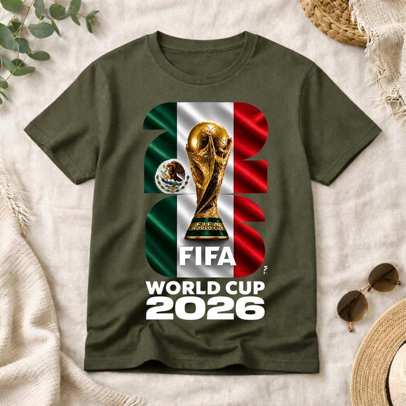Pu&ograve; includere: Una t-shirt verde oliva con una grande grafica del trofeo della Coppa del Mondo FIFA 2026 centrata sulla bandiera messicana. Il design include il testo 2026, FIFA e WORLD CUP 2026 in caratteri bianchi, con lo stemma messicano visibile.