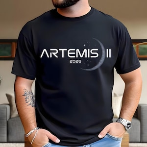 Puede incluir: Camiseta negra con el texto blanco "ARTEMIS II" y el año "2026". Un gráfico de media luna con estrellas está a la derecha del texto. La camiseta es de manga corta.
