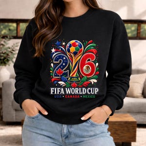 May include: Black crewneck sweatshirt with a colorful graphic for the FIFA World Cup 2026. The design features the numbers 2 and 6, a soccer ball, and the flags of USA, Canada, and Mexico. The text "FIFA WORLD CUP" is displayed below.