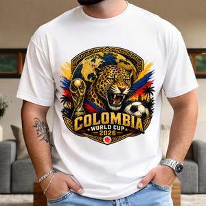 May include: White t-shirt featuring a vibrant graphic design for the Colombia World Cup 2026. The design includes a jaguar, a soccer ball, the FIFA trophy, and the text "COLOMBIA WORLD CUP 2026" in gold and red.