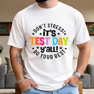 Puede incluir: Camiseta blanca con el texto "DON'T STRESS! IT'S TEST DAY y'all! DO YOUR BEST" en un diseño circular. El texto es de varios colores, incluyendo naranja, rosa, azul y verde. La camiseta está siendo usada.