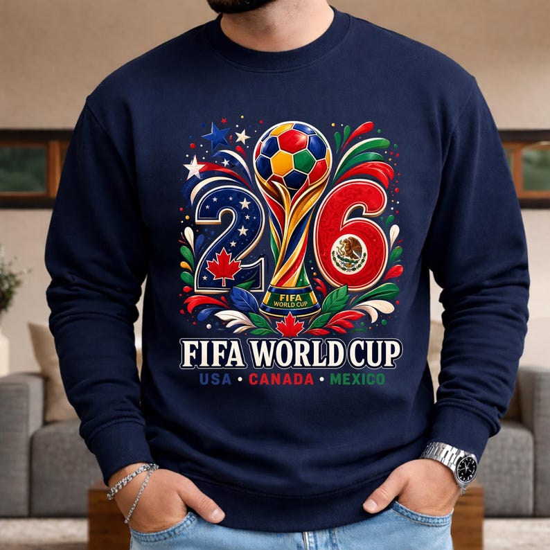 May include: Navy blue sweatshirt featuring a colorful graphic for the FIFA World Cup 2026. The design includes a trophy, soccer ball, and the numbers 2 and 6. The text "FIFA WORLD CUP" is displayed below, with the USA, Canada, and Mexico flags.
