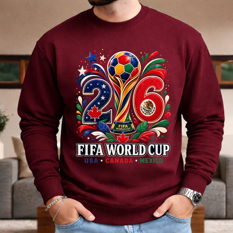May include: Burgundy sweatshirt featuring a colorful graphic for the 2026 FIFA World Cup. The design includes a trophy, a soccer ball, and the numbers 2 and 6. The text "FIFA WORLD CUP" is displayed below, with "USA CANADA MEXICO" underneath.