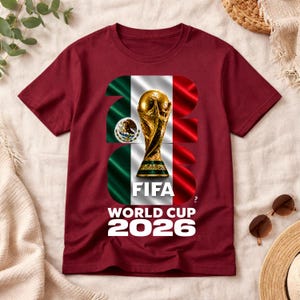 Pu&ograve; includere: Una t-shirt a maniche corte color bordeaux con una grande grafica del trofeo della Coppa del Mondo FIFA 2026 centrata sulla bandiera messicana. Il design include il testo FIFA WORLD CUP 2026 in lettere bianche, con il numero 2026 sullo sfondo.