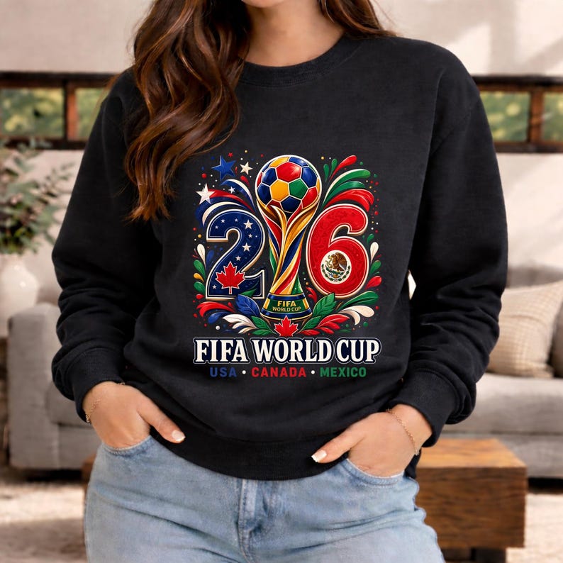 May include: Black sweatshirt featuring a colorful graphic for the 2026 FIFA World Cup. The design includes a soccer ball, the numbers 2 and 6, and the text "FIFA WORLD CUP USA CANADA MEXICO". The sweatshirt has a crew neck.