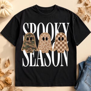 Peut inclure: Un t-shirt noir à manches courtes avec le texte blanc SPOOKY SEASON en grande police. Trois graphismes en forme de fantôme sont centrés sur le t-shirt, chacun avec un motif différent : un camouflage vert et marron, un imprimé léopard beige et un motif à damier marron.