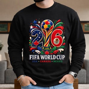 May include: Black sweatshirt featuring a colorful graphic for the 2026 FIFA World Cup. The design includes a trophy, soccer ball, and the numbers 2 and 6. The text "FIFA WORLD CUP" is displayed below, with "USA CANADA MEXICO" underneath.