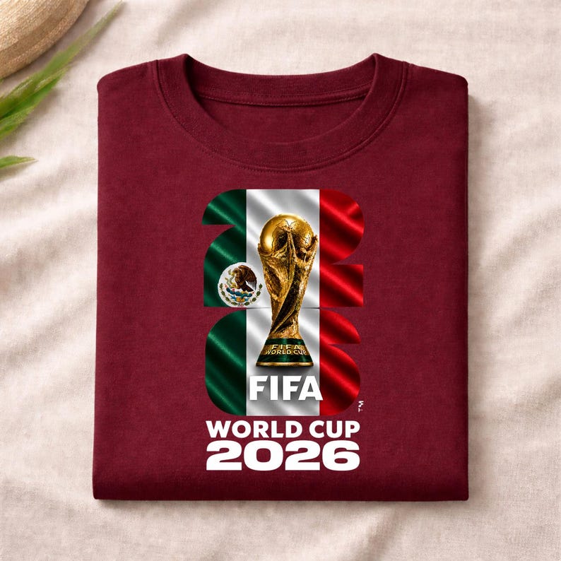 Pu&ograve; includere: Una t-shirt bordeaux a girocollo con una grafica del trofeo della Coppa del Mondo FIFA 2026 centrata su una bandiera messicana ondulata. Il design incorpora il numero 2026 in grandi caratteri bianchi, con FIFA e World Cup 2026 stampati sotto.