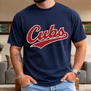 May include: Navy blue t-shirt with the word "Cubs" in distressed red script and a white outline. The short-sleeved crew neck shirt is suitable for casual wear. The design is centrally placed on the chest.