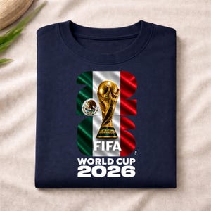 Pu&ograve; includere: Una t-shirt blu navy con una grande grafica del trofeo della Coppa del Mondo FIFA 2026 centrata nel numero 2026. Il disegno incorpora i colori della bandiera messicana verde, bianco e rosso, insieme allo stemma nazionale del Messico.