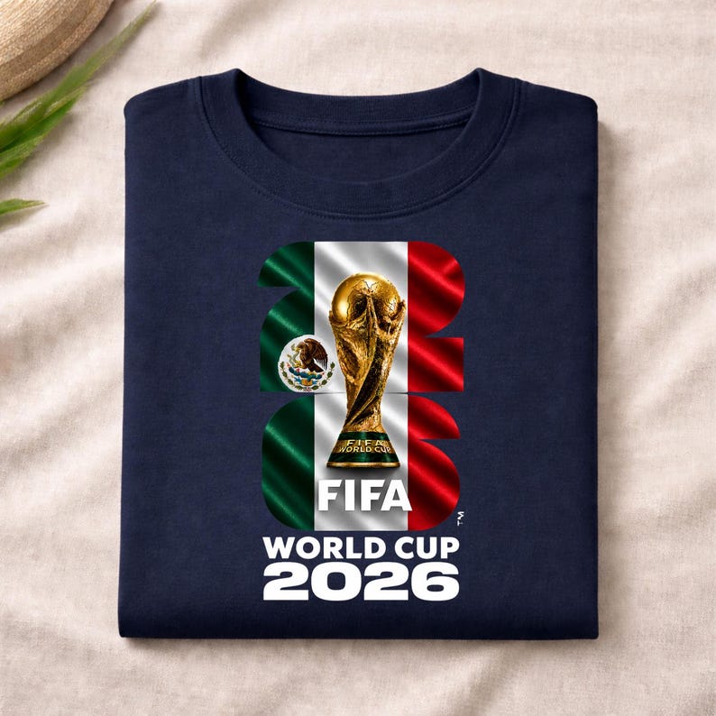 Pu&ograve; includere: Una t-shirt blu navy con una grande grafica del trofeo della Coppa del Mondo FIFA 2026 centrata nel numero 2026. Il disegno incorpora i colori della bandiera messicana verde, bianco e rosso, insieme allo stemma nazionale del Messico.