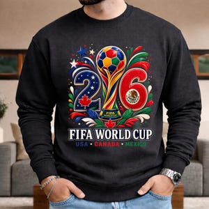 May include: Dark gray sweatshirt featuring a colorful graphic for the 2026 FIFA World Cup. The design includes the numbers 2 and 6, a trophy, and the flags of USA, Canada, and Mexico. The words "FIFA WORLD CUP" are also displayed.